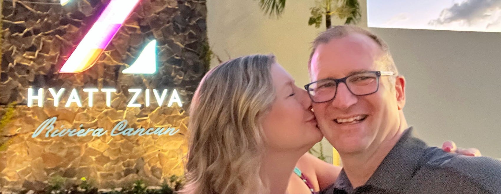Zuzu kissing Ross in front of the sign for Hyatt Ziva Riviera Cancun All Inclusive resort