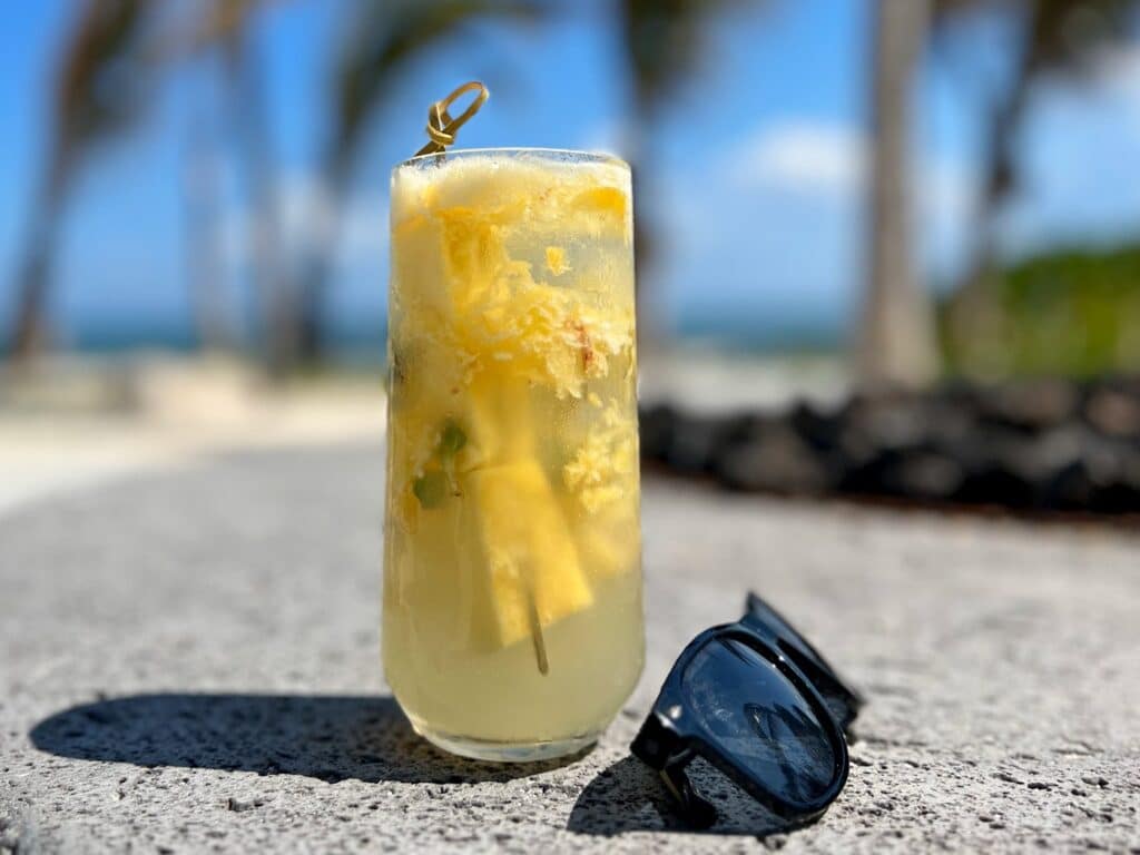 Pineapple Mojito and sunglasses on a grey rock with palm trees in the background
